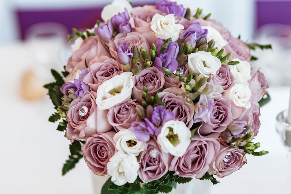 A close-up image of a floral bouquet featuring soft pink roses, white lisianthus, and purple freesia arranged together with lush green foliage. The flowers are fresh, with delicate petals showcasing s