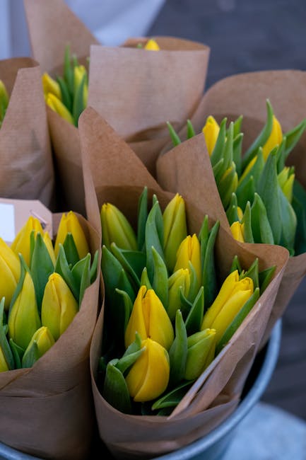 A close-up view of multiple bouquets containing fresh yellow tulips with green leaves, arranged in brown kraft paper wrapping. The tulips display vibrant yellow petals, some partially open, with smoot