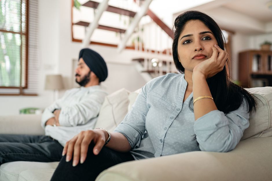 A young woman with long black hair, wearing a light blue blouse and a gold bracelet, is sitting on a white sofa with her head resting on her hand, looking thoughtful or bored. Behind her, a man wearin