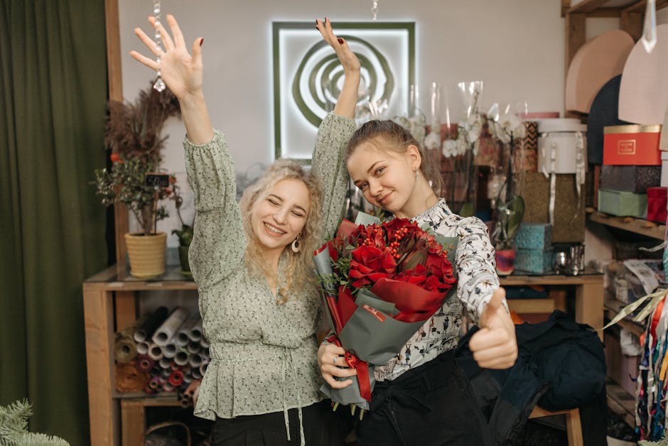 Two young women inside a floral shop, one holding a lush bouquet of red roses wrapped in grey paper with a red ribbon, and the other smiling with her arms raised in celebration. The woman with the bou