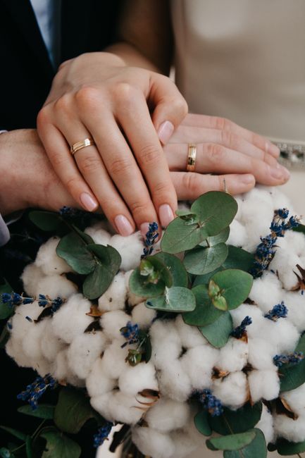 Close-up of a couple's hands gently resting on a bridal bouquet consisting of fluffy white cotton blossoms and sprigs of small blue flowers, accented with broad, round eucalyptus leaves. The bride and