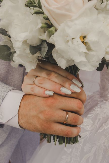 A close-up of a bride and groom's hands holding a bouquet of fresh, predominantly white and cream flowers, including lisianthus and possibly roses, accented with green foliage. The bride's hand, featu